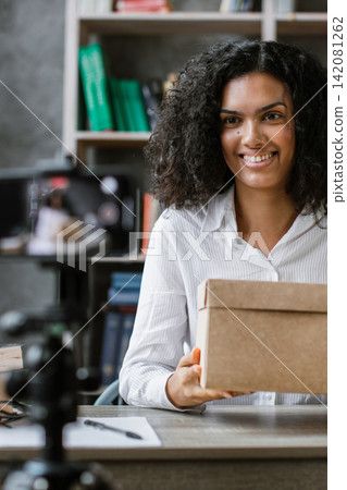 A young woman smiles while holding a cardboard box in front of a smartphone recording her content 142081262