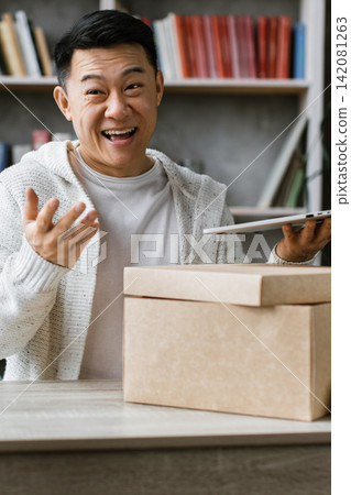 A cheerful Asian man records himself unboxing a package while holding a laptop, engaging his audience with enthusiasm A cheerful Asian man records himself unboxing a package while holding a laptop, engaging his audience with enthusiasm 142081263