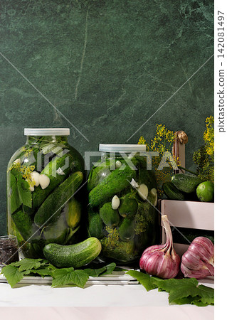 Cucumbers in a basket with dill, garlic, pickling leaves, and pickled cucumbers in jars on a kitchen counter. Harvest concept. Homemade pickle recipe and ingredients. Home canning concept. Cucumbers in a basket with dill, garlic, pickling leaves, and pickled cucumbers in jars on a kitchen counter. Harvest concept. Homemade pickle recipe and ingredients. Home canning concept. 142081497