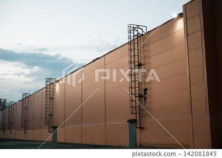 A large commercial or industrial building photographed at dusk, featuring a long peach-colored exterior facade, multiple metal emergency access ladders, service doors and exterior security lighting A large commercial or industrial building photographed at dusk, featuring a long peach-colored exterior facade, multiple metal emergency access ladders, service doors and exterior security lighting 142081845