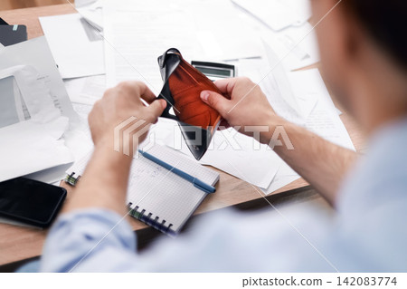 Man holding an empty wallet over a desk with unpaid bills. He faces financial problems, rising energy prices, and household expenses due to inflation and an economic crisis. Man holding an empty wallet over a desk with unpaid bills. He faces financial problems, rising energy prices, and household expenses due to inflation and an economic crisis. 142083774