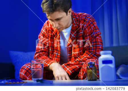 An unwell sick man sits at home in pajamas, suffering from a seasonal cold and flu infection. He looks at pills and medicine for respiratory illness treatment and recovery. An unwell sick man sits at home in pajamas, suffering from a seasonal cold and flu infection. He looks at pills and medicine for respiratory illness treatment and recovery. 142083779