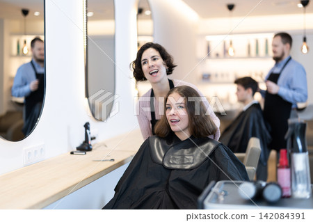 Young woman barbershop employee stands next to female client, discussing details of haircut Young woman barbershop employee stands next to female client, discussing details of haircut 142084391