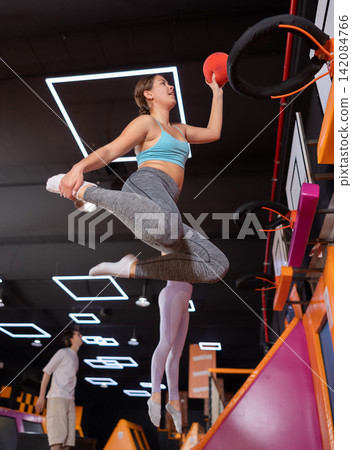 Cheerful young European female in sportswear playing basketball while bouncing on colorful trampolines indoors in leisure sports center 142084766