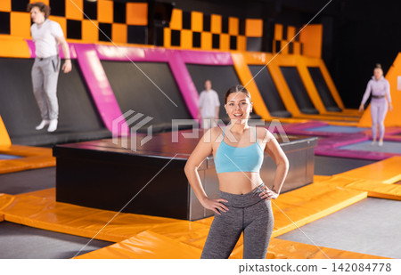 Happy young European female in blue top and gray leggings posing on trampolines during active weekend free time in sports center Happy young European female in blue top and gray leggings posing on trampolines during active weekend free time in sports center 142084778