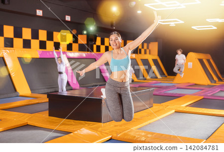 Excited young woman in blue top and gray leggings having great time while jumping on colorful trampoline in game club Excited young woman in blue top and gray leggings having great time while jumping on colorful trampoline in game club 142084781