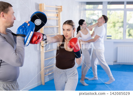 Woman wearing boxing gloves exercise and punch to pads for boxing. Woman with trainer at gym Woman wearing boxing gloves exercise and punch to pads for boxing. Woman with trainer at gym 142085029