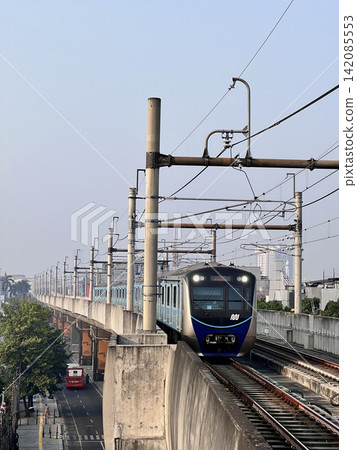 A modern electric commuter train traveling along an elevated railway in a busy city environment, approaching on parallel tracks beneath a network of overhead electrical wires and supporting poles. 142085553