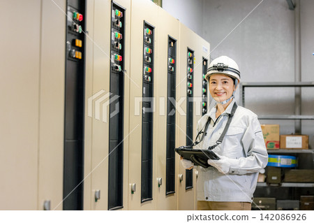 A woman in work clothes holding a tablet in front of an electronic control panel. A woman in work clothes holding a tablet in front of an electronic control panel. 142086926