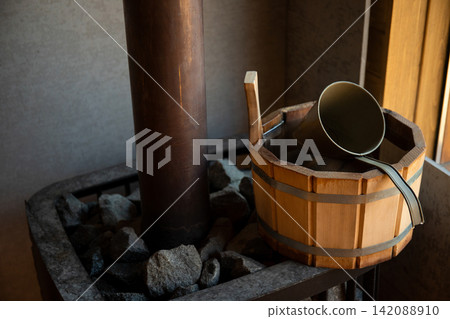 Wooden bucket and metal ladle sit beside a stone heater in a tranquil sauna setting Wooden bucket and metal ladle sit beside a stone heater in a tranquil sauna setting 142088910
