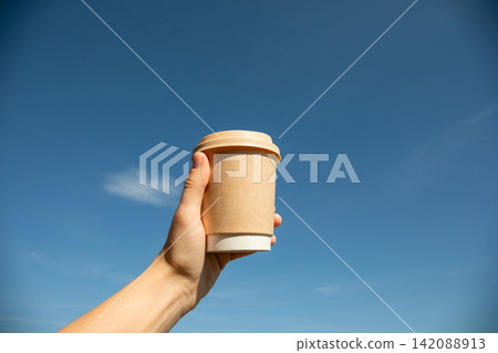 Hand displays sustainable coffee cup under sunny sky. Eco-friendly lifestyle concept Hand displays sustainable coffee cup under sunny sky. Eco-friendly lifestyle concept 142088913