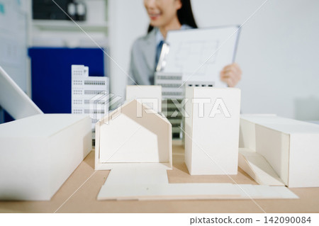 Female architect designing a sustainable urban building project model with solar panels, analyzing blue-print plans in office. Female architect designing a sustainable urban building project model with solar panels, analyzing blue-print plans in office. 142090084