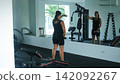 Young woman in black sportswear performing dumbbell front raise exercise with arms extended forward in a bright modern gym. Front deltoid training concept 142092267