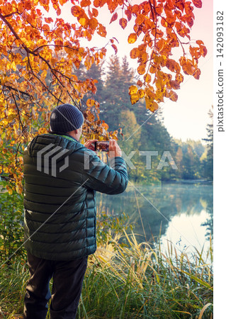 A man stands on the picturesque lakeshore in the evening. The man takes a photo of the lake with a mobile phone A man stands on the picturesque lakeshore in the evening. The man takes a photo of the lake with a mobile phone 142093182
