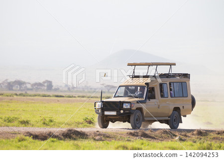 A beige 4x4 safari truck with a raised pop-up roof driving along a dirt road through the open grassy savanna plains of an African national park during a game drive excursion. A beige 4x4 safari truck with a raised pop-up roof driving along a dirt road through the open grassy savanna plains of an African national park during a game drive excursion. 142094253