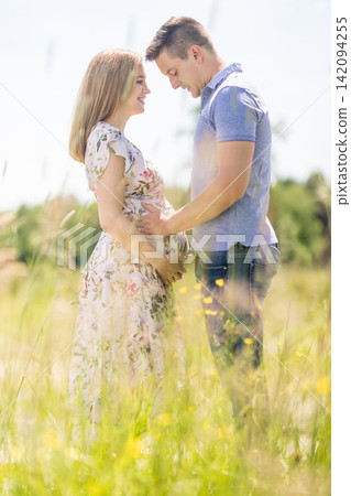 Side view of a happy pregnant couple embracing gently and holding the belly in a sunlit grassy field during summer. Romantic maternity concept of love, family preparation, and new life. Side view of a happy pregnant couple embracing gently and holding the belly in a sunlit grassy field during summer. Romantic maternity concept of love, family preparation, and new life. 142094255