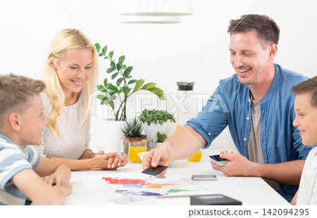 Happy family playing a world map educational board game together around table. Father placing card on world map board game while family watches and smiles. Happy family playing a world map educational board game together around table. Father placing card on world map board game while family watches and smiles. 142094295