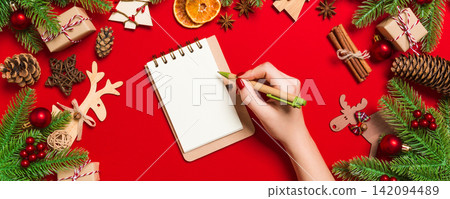 Top view of female hand making some notes in noteebok on colorful background. New Year decorations and toys. Christmas time concept Top view of female hand making some notes in noteebok on colorful background. New Year decorations and toys. Christmas time concept 142094489