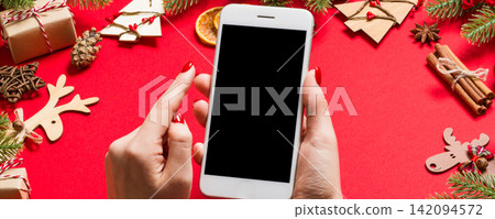 Top view of phone in female hand on festive red background. Christmas decorations. New Year time holiday. Mockup Top view of phone in female hand on festive red background. Christmas decorations. New Year time holiday. Mockup 142094572