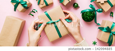 Woman is packing surprise gift for Christmas holidays, sitting at workplace with craft tools, top view. Woman wrapping Christmas gift at colored table Woman is packing surprise gift for Christmas holidays, sitting at workplace with craft tools, top view. Woman wrapping Christmas gift at colored table 142094885