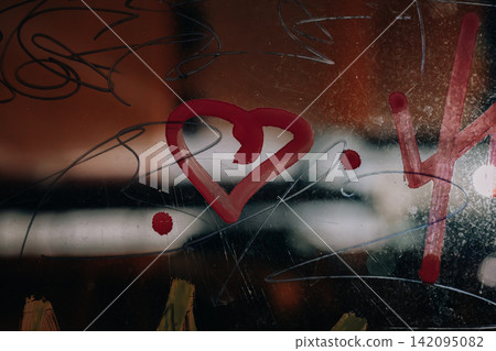 Artistic close-up of a red heart symbol painted on a glass surface against a dark nighttime background with blurred city lights and reflections. Scratches, abstract lines and glowing bokeh at night. Artistic close-up of a red heart symbol painted on a glass surface against a dark nighttime background with blurred city lights and reflections. Scratches, abstract lines and glowing bokeh at night. 142095082