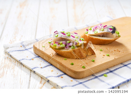 Slices of baguette with pieces marinated herring fillet with with red onions and pickles on table. Slices of baguette with pieces marinated herring fillet with with red onions and pickles on table. 142095194