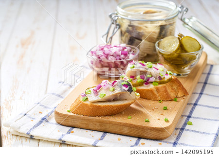 Slices of baguette with pieces marinated herring fillet with with red onions and pickles on table. Slices of baguette with pieces marinated herring fillet with with red onions and pickles on table. 142095195