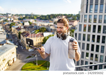 A bearded man with headphones around his neck records himself on a smartphone attached to a selfie stick while holding a large water bottle on a rooftop overlooking a city A bearded man with headphones around his neck records himself on a smartphone attached to a selfie stick while holding a large water bottle on a rooftop overlooking a city 142095692
