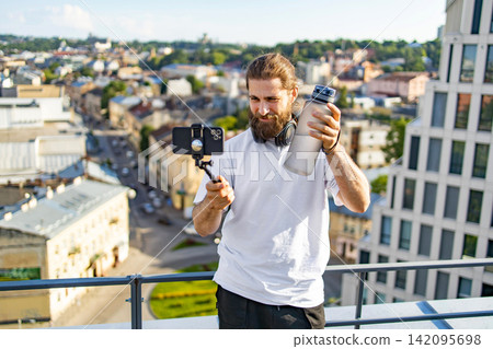 A man with a beard and headphones films himself with a smartphone on a selfie stick while holding a water bottle on a rooftop overlooking a city A man with a beard and headphones films himself with a smartphone on a selfie stick while holding a water bottle on a rooftop overlooking a city 142095698