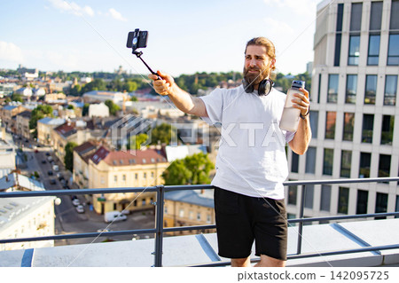 A bearded man in athletic wear holds a water bottle and a selfie stick while recording a video on a rooftop overlooking a city A bearded man in athletic wear holds a water bottle and a selfie stick while recording a video on a rooftop overlooking a city 142095725