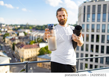 A man with a beard and bun hairstyle holds a water bottle and a rolled-up yoga mat on a rooftop overlooking a city skyline A man with a beard and bun hairstyle holds a water bottle and a rolled-up yoga mat on a rooftop overlooking a city skyline 142095831