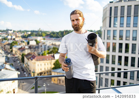 A bearded man in a white t-shirt stands on a rooftop holding a yoga mat and a water bottle with a city view behind him A bearded man in a white t-shirt stands on a rooftop holding a yoga mat and a water bottle with a city view behind him 142095833