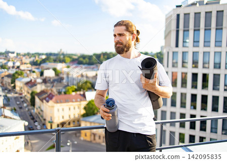 A bearded man with his hair tied back holds a yoga mat and water bottle while standing on a rooftop overlooking a city A bearded man with his hair tied back holds a yoga mat and water bottle while standing on a rooftop overlooking a city 142095835