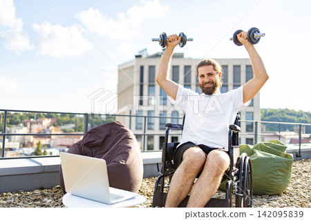 A smiling man with a beard sits in a wheelchair on a rooftop terrace, lifting dumbbells overhead while looking at the camera A smiling man with a beard sits in a wheelchair on a rooftop terrace, lifting dumbbells overhead while looking at the camera 142095839
