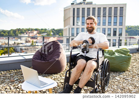 A smiling man in a wheelchair is lifting dumbbells on a rooftop terrace with a city view, accompanied by beanbag chairs and a laptop 142095902