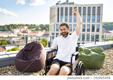 A man with a beard and long hair in a wheelchair raises his fist in triumph on a rooftop terrace with a city view A man with a beard and long hair in a wheelchair raises his fist in triumph on a rooftop terrace with a city view 142095949