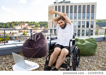 A smiling man in a wheelchair stretches his arms on a rooftop patio with beanbag chairs and a laptop nearby, overlooking a city skyline A smiling man in a wheelchair stretches his arms on a rooftop patio with beanbag chairs and a laptop nearby, overlooking a city skyline 142095956