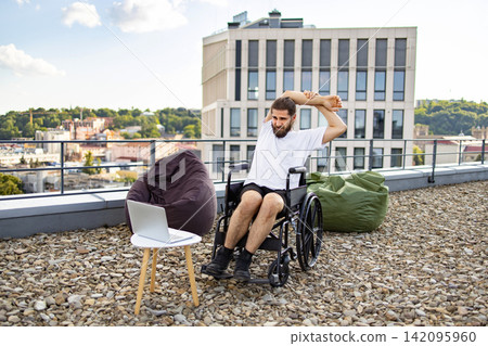 A man in a wheelchair stretches his arms on a rooftop terrace with a city view, beanbags, and a laptop nearby 142095960