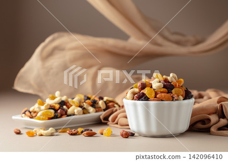 The mix of various nuts and raisins in a white bowl on a beige background. The mix of various nuts and raisins in a white bowl on a beige background. 142096410