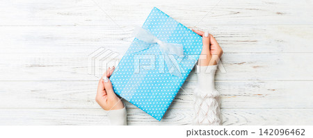 Woman arms holding gift box with colored ribbon on white rustic wooden table background, top view and copy space for you design Woman arms holding gift box with colored ribbon on white rustic wooden table background, top view and copy space for you design 142096462