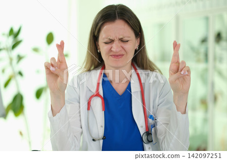 Female doctor with eyes closed tightly raises crossed fingers Female doctor with eyes closed tightly raises crossed fingers 142097251