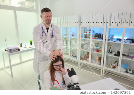 Doctor supervising young student with brain model in bright medical laboratory, healthcare education scene with microscope, lab coats, shelves, clinical study tools, professional mentoring Doctor supervising young student with brain model in bright medical laboratory, healthcare education scene with microscope, lab coats, shelves, clinical study tools, professional mentoring 142097801
