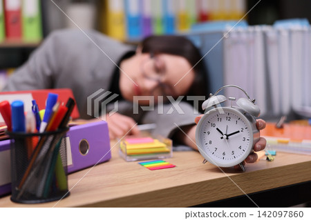 Exhausted businesswoman resting head on desk while holding ringing alarm clock among pens, folders, notes, and colorful stationery in cluttered office, showing overtime stress and missed rest Exhausted businesswoman resting head on desk while holding ringing alarm clock among pens, folders, notes, and colorful stationery in cluttered office, showing overtime stress and missed rest 142097860