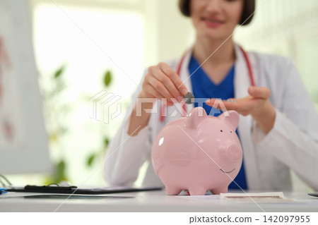 Woman doctor puts coin into piggy bank for save at clinic desk Woman doctor puts coin into piggy bank for save at clinic desk 142097955