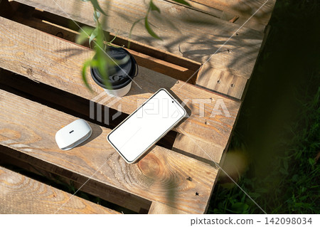 Smartphone, wireless earbuds and reusable coffee cup on rustic wooden table outdoors. Smartphone, wireless earbuds and reusable coffee cup on rustic wooden table outdoors. 142098034