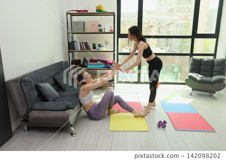 Two women practicing assisted stretching and balance exercises in bright apartment living room with mats, sofa, shelves, large windows, and natural daylight, with women, exercise, stretching 142098202