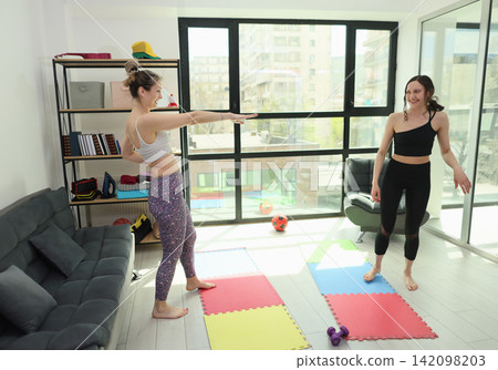 Two women smiling during home fitness routine in bright modern living room with yoga mats, exercise props, large windows, city view, natural daylight and relaxed wellness mood Two women smiling during home fitness routine in bright modern living room with yoga mats, exercise props, large windows, city view, natural daylight and relaxed wellness mood 142098203