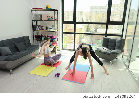 Two women practicing yoga and stretching on colorful mats in bright modern living room with large windows, city view, shelving, fitness props, relaxed home wellness routine Two women practicing yoga and stretching on colorful mats in bright modern living room with large windows, city view, shelving, fitness props, relaxed home wellness routine 142098204