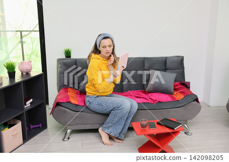 Woman in yellow hoodie sits on modern sofa and checks her face in handheld mirror inside bright living room with pink accents, casual home routine, relaxed morning preparation, indoor lifestyle scene Woman in yellow hoodie sits on modern sofa and checks her face in handheld mirror inside bright living room with pink accents, casual home routine, relaxed morning preparation, indoor lifestyle scene 142098205