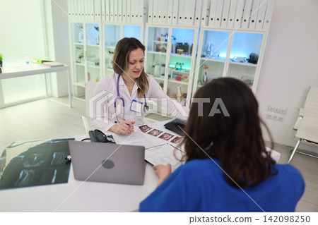 Smiling doctor reviewing medical forms with nurse during consultation in modern clinic office with laptop, clipboard, x ray image, white cabinets, bright professional healthcare setting Smiling doctor reviewing medical forms with nurse during consultation in modern clinic office with laptop, clipboard, x ray image, white cabinets, bright professional healthcare setting 142098250